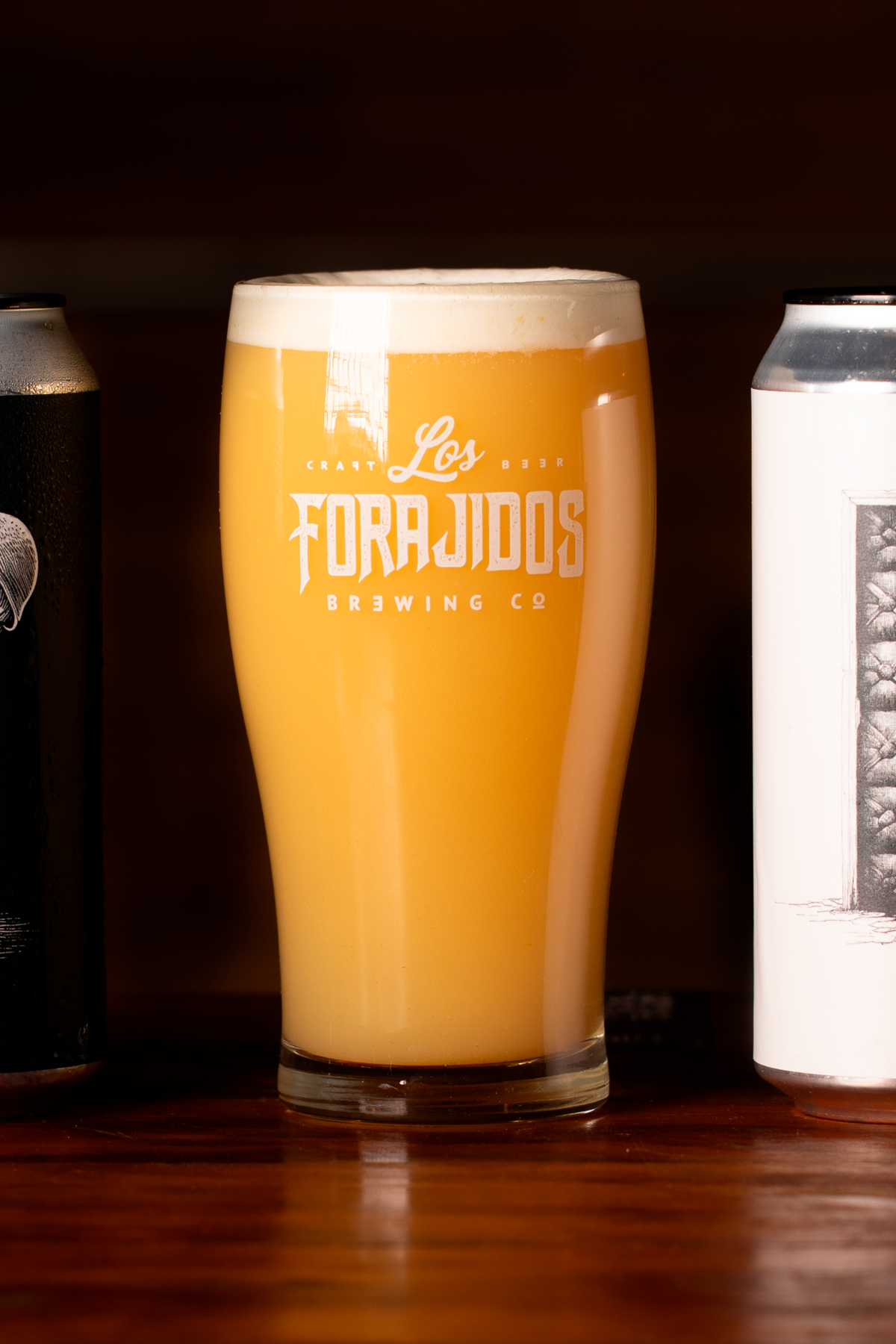 Copo de hazy IPA Los Forajidos entre latas sobre balcão de madeira